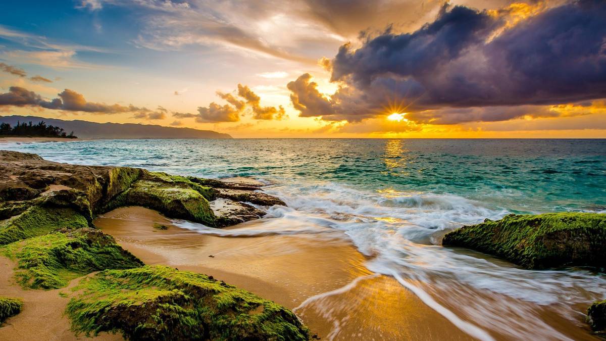 Blue green ocean with sand and algae covered rocks in foreground, big grey clouds and low sun in background at baldwin beach maui hawaii